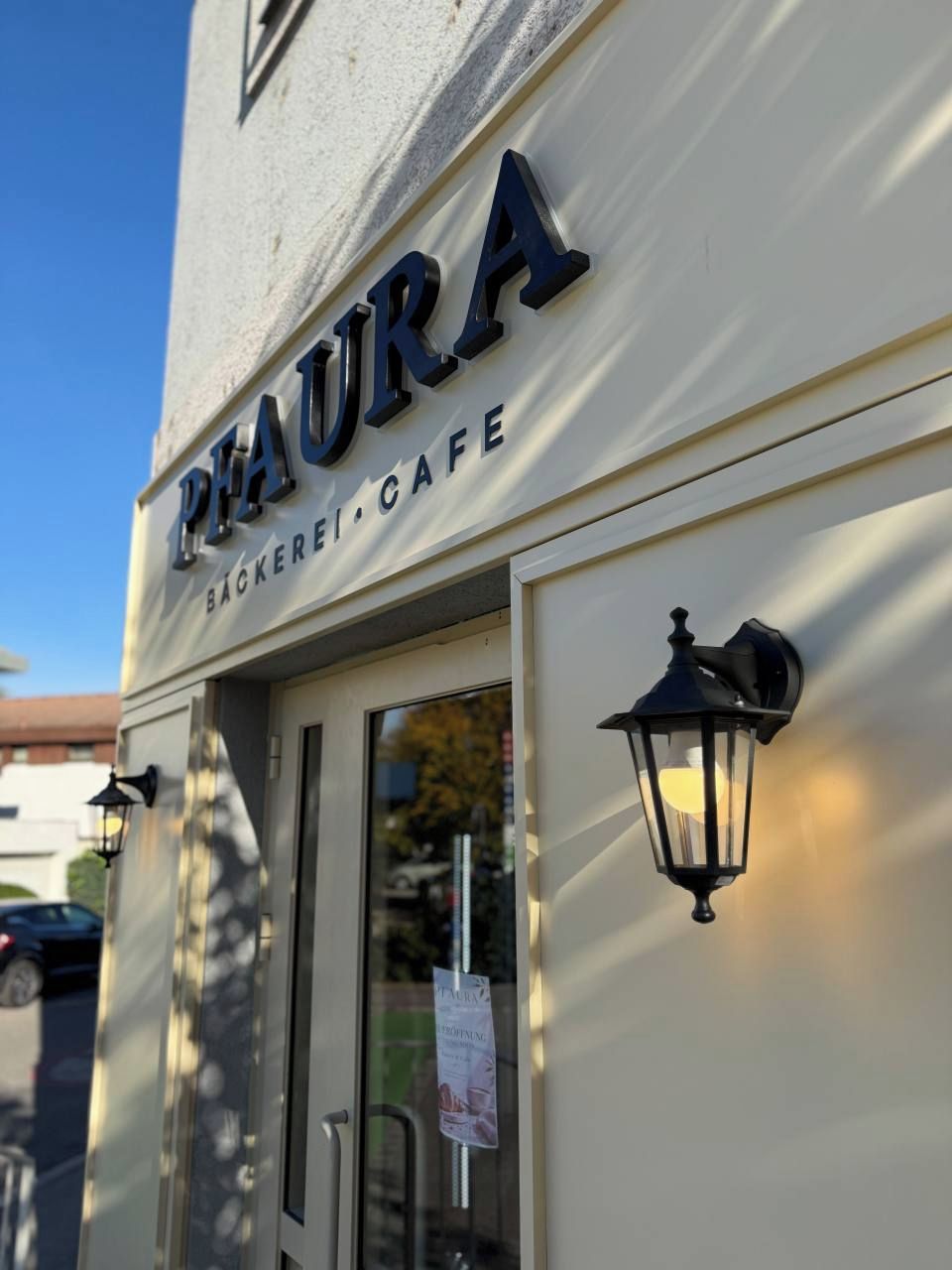Een gebouw met een bord waarop 'PHAURA BÄCKEREI CAFE' staat en een zwart, aan de muur bevestigd lampje.