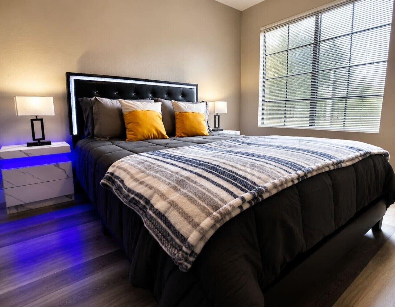 A bedroom with a large bed, two bedside tables, a window with blinds, and a nightstand with a lamp.