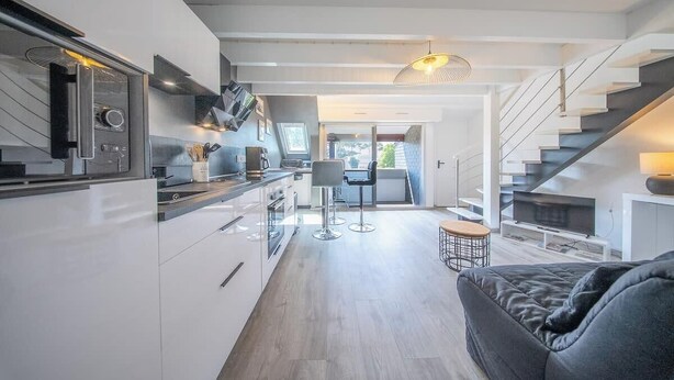 Una cocina moderna con armarios blancos, una zona de comedor con un taburete de bar, un sofá y una escalera que conduce a un balcón.