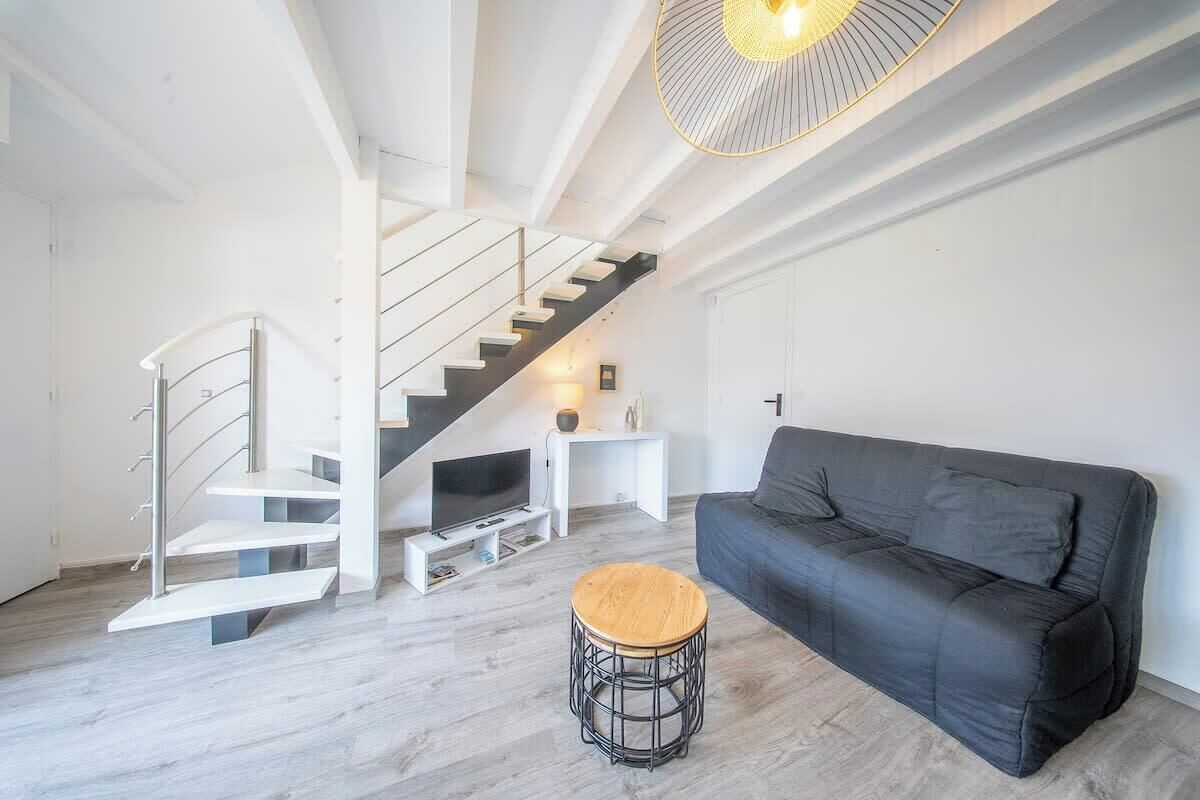 Een moderne woonkamer met een zwarte bank, een houten kruk, een witte trap en een televisie op een witte dressoir.