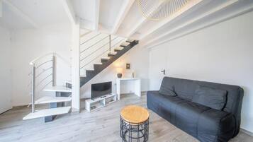 Een moderne woonkamer met een zwarte bank, een houten kruk, een witte trap en een televisie op een witte dressoir.