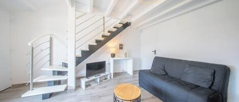 A modern living room with a black sofa, a wooden stool, a white staircase, and a television on a white stand.