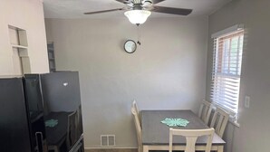 Une cuisine de taille réduite comprenant un réfrigérateur, une table à manger, des chaises, un ventilateur de plafond et une fenêtre avec des stores.