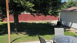 Une terrasse extérieure avec une table ronde en verre et des chaises en métal, un poteau en bois et, au fond, une clôture rouge.