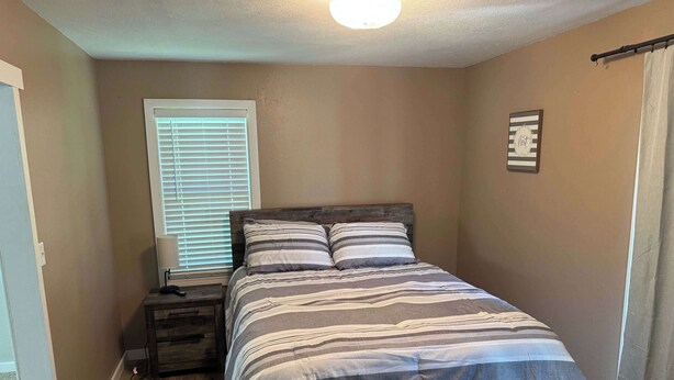 Une chambre à coucher avec un lit en bois, une table de chevet, une lampe, une fenêtre avec des stores et un élément de décoration fixé au mur.