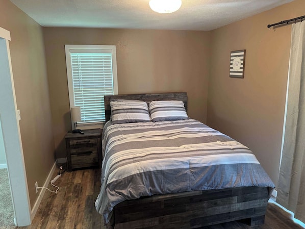 A bedroom with a wooden bed, a nightstand, a lamp, a window with blinds, and a wall-mounted decoration.