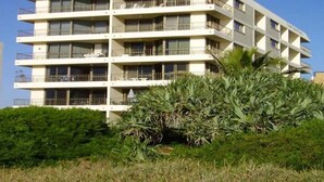 Ein mehrstöckiges Hotel mit Balkonen, einer flachen Dachterrasse und einem üppigen Grünstrauch im Vordergrund.