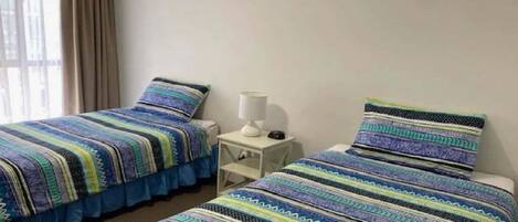 Dos camas individuales con ropa de cama a rayas, una mesita de noche con lámpara y una ventana con cortinas.