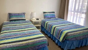 Una habitación con dos camas, cada una con una colcha estampada, una mesita de noche con lámpara y una ventana con cortinas.
