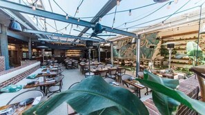 Un espacio exterior para comer con mesas y sillas, un muro de ladrillo y luces colgantes.