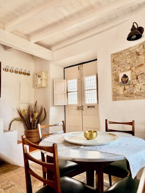 Uma sala de jantar com uma mesa redonda, cadeiras, um vaso com plantas secas, uma luminária na parede e uma parede de pedra com um quadro.