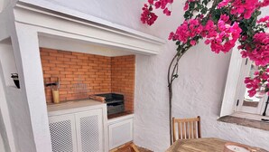 Eine Terrasse mit einer Backsteinmauer, einem Grill mit weißem Rahmen, einem Holztisch mit Kissen und pinkfarbenen Bougainvillea-Blüten.