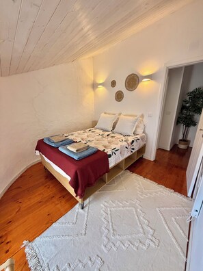 Une chambre avec un plafond en bois, un lit recouvert d’une couvre-lit à motifs floraux, un plaid rouge et un tapis blanc.