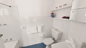 Et badeværelse med hvide fliser, toilet, vask og brusebad. Der er håndklæder hængende på en holder, og en hylde med toiletartikler.