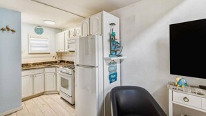 A compact kitchen with white cabinets, a stove, a microwave, and a refrigerator. There is a small shelf with decorative items and a clock on the wall. - Beachfront location, steps from the boardwalk, Fri night fireworks & more! (North Wildwood)