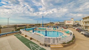 A pool area with lounge chairs and umbrellas, a parking lot, and a building with 'AMERICAN' signage. - Beachfront location, steps from the boardwalk, Fri night fireworks & more! (North Wildwood)