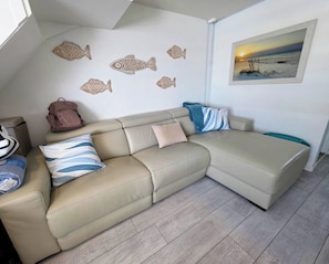 A living room with a beige sectional sofa, decorative fish wall art, a framed landscape painting, and a blue-striped pillow. - Beachfront location, steps from the boardwalk, Fri night fireworks & more! (North Wildwood)