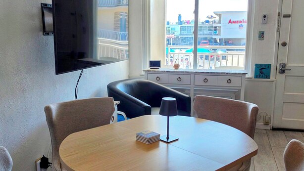 A room with a round wooden table, four upholstered chairs, a flat-screen TV mounted on the wall, and a window with a view of a beach and buildings. - Beachfront location, steps from the boardwalk, Fri night fireworks & more! (North Wildwood)