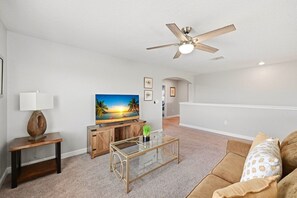 A living room with a ceiling fan, a television, a coffee table, a couch, and a painting on the wall. - Pool Home w/ Spa & Game Room · 15 Min to Disney · 5BR Sleeps 10 (Four Corners)