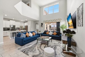 A spacious living area with two sofas, a coffee table, and a rug. There is a kitchen in the background, a dining area, and a large window. - Renovated 5BR Pool Home · Game Room · 15 Min to Disney · Sleeps 10 (Four Corners)