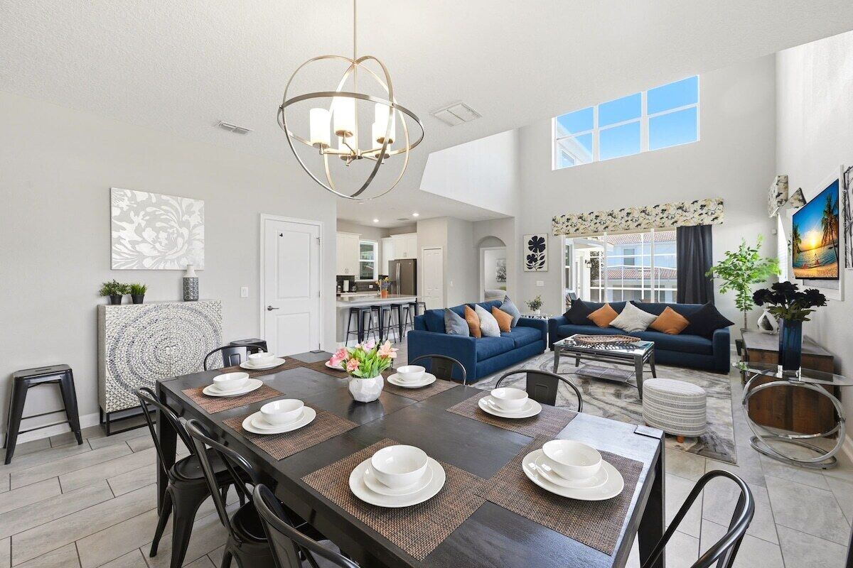 A modern living room with a dark wooden dining table set for four, a blue sofa, and a large window with a view.