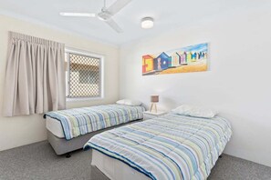Ein Schlafzimmer mit zwei Einzelbetten, einem fensterbelaubten Fenster, einem Deckenventilator und einem Wandbild.