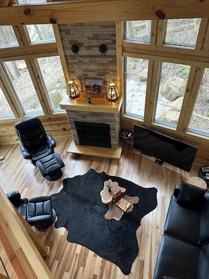 Um quarto aconchegante com lareira de pedra, poltronas de couro, tapete preto e janelas grandes.