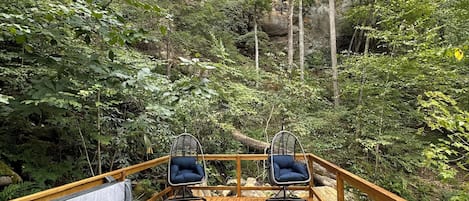 A wooden deck with a hot tub and two chairs overlooking a forested cliff.