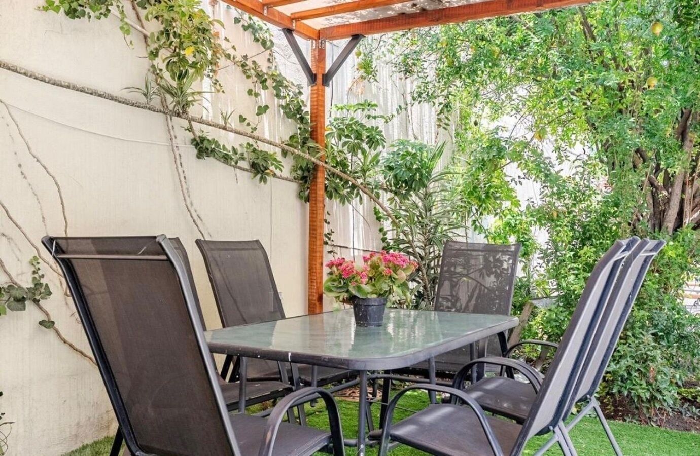Une terrasse extérieure avec une table en verre et des chaises en métal, entourée de verdure et d’une pergola en bois.