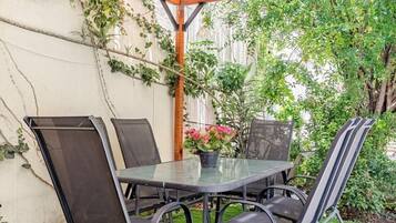 Une terrasse extérieure avec une table en verre et des chaises en métal, entourée de verdure et d’une pergola en bois.
