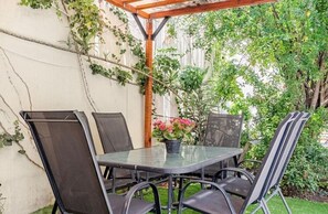 Un patio al aire libre con una mesa de vidrio y sillas de metal, rodeado de follaje y una pérgola de madera.