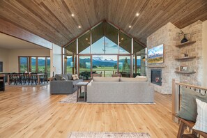 A spacious living area with a large window overlooking a mountainous landscape, a fireplace, and a dining area with wooden furniture. - Luxury Retreat, Teton Views, Sleeps 16+, Hot Tub, Fire Pit (Tetonia)