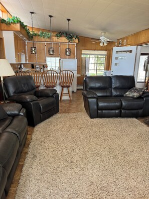 A living room with black leather sofas, a wooden dining table with chairs, a kitchen area with wooden cabinets and a refrigerator, and a ceiling fan. - Spacious 4-bedroom house on all sports 8 Pt Lake in wonderful Lake with AC (Lake)