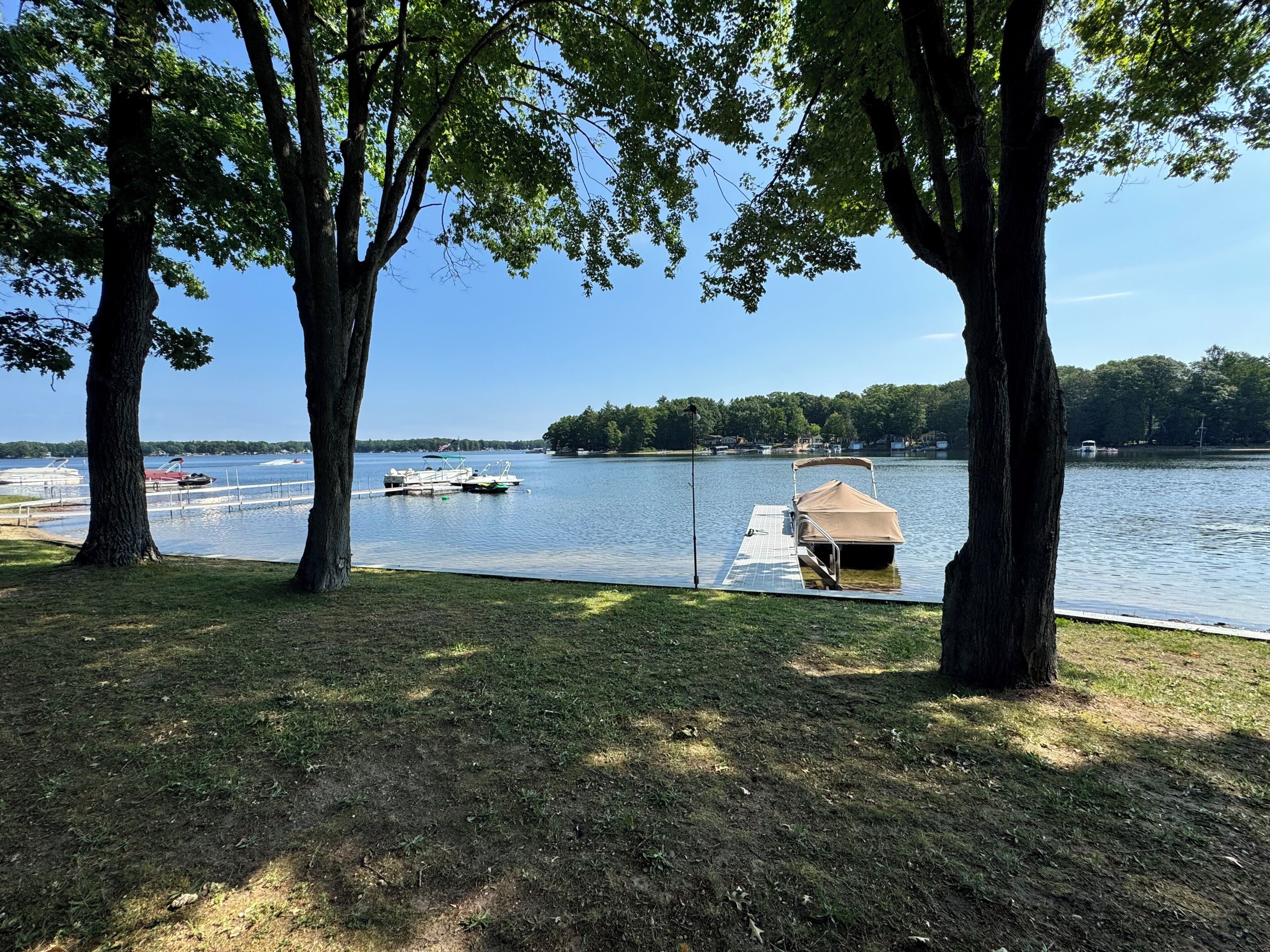 A lakeside view with docked boats, a covered boat, and a grassy area with trees.