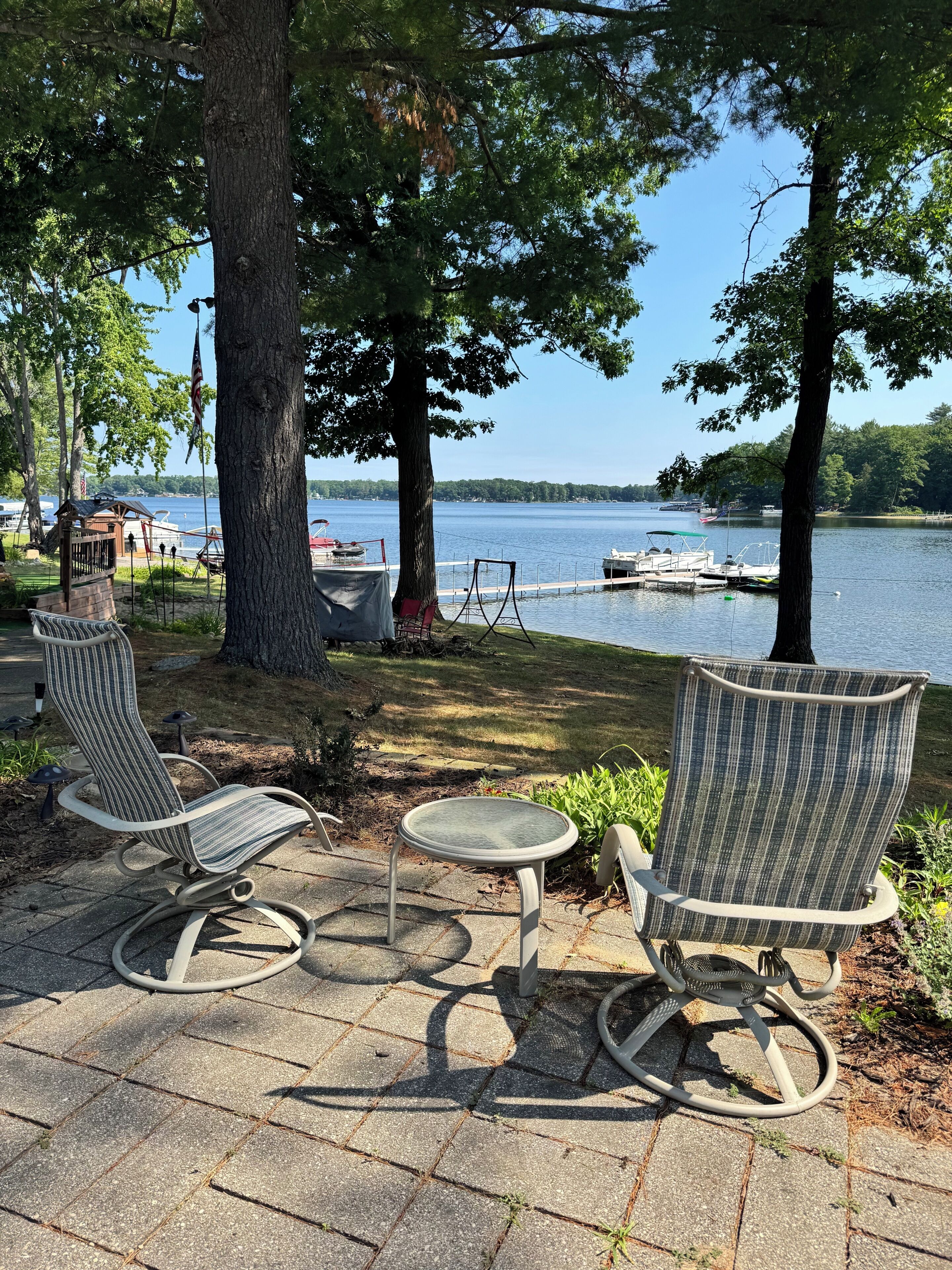 A lakeside patio with two rocking chairs and a small table, overlooking a dock with boats.