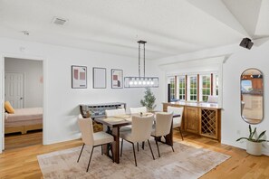 A dining area with a table, chairs, a hanging light fixture, and a view of the kitchen through an open door.