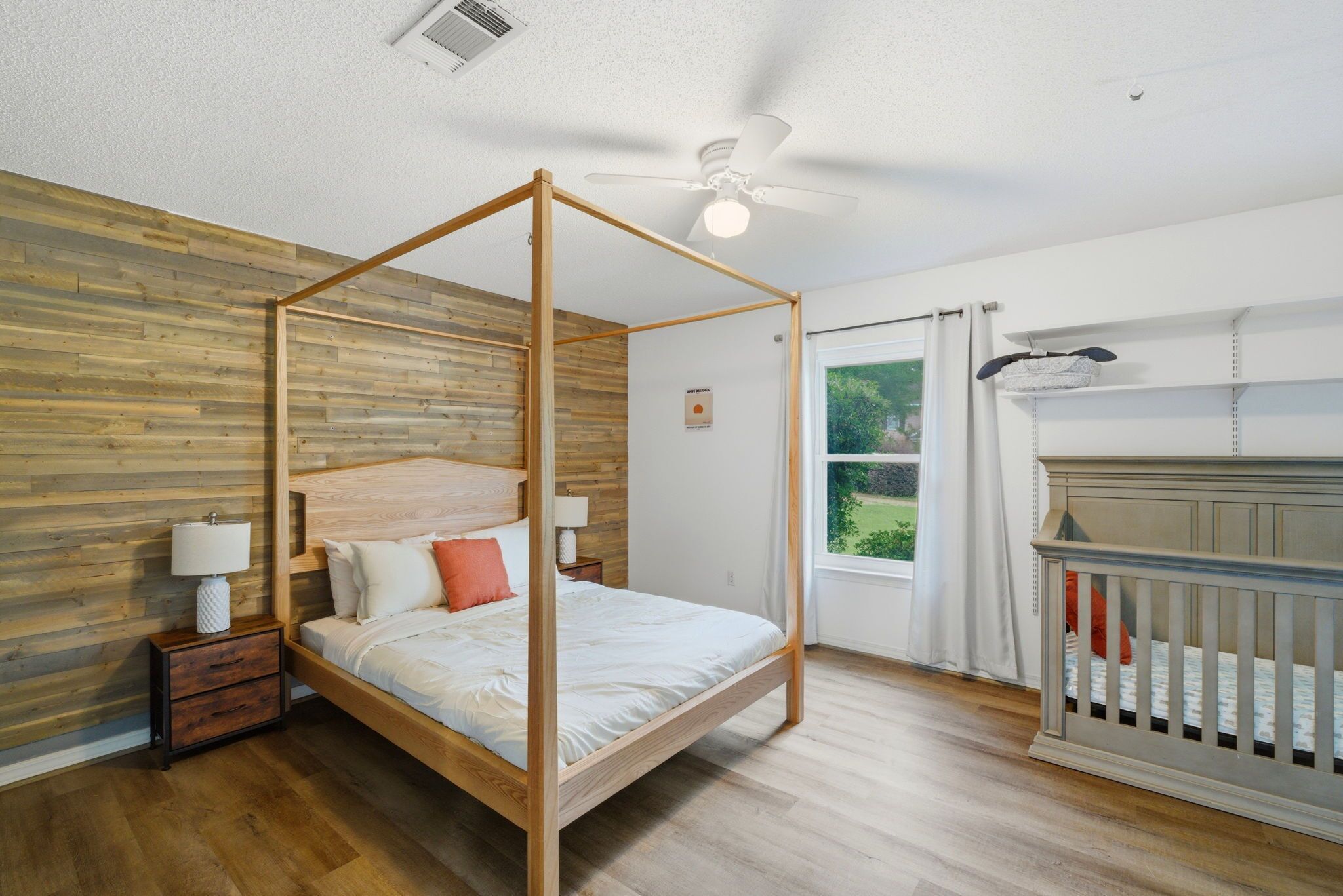 A four-poster bed with a wooden headboard, a nightstand with a lamp, a crib, and a view of greenery outside.