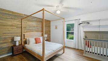 A four-poster bed with a wooden headboard, a nightstand with a lamp, a crib, and a view of greenery outside.