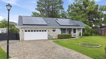 A single-story house with a garage, solar panels on the roof, a brick driveway, and a well-maintained lawn.