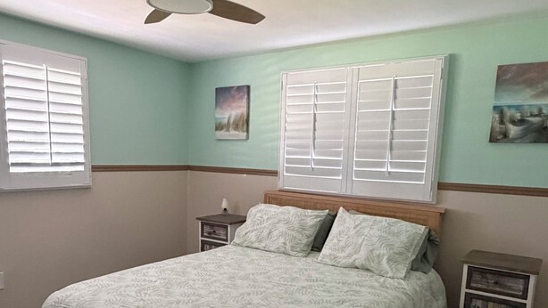 Quarto com uma cama, ventoinha no teto, duas janelas com cortinas e uma mesa de cabeceira.