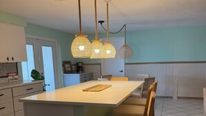 Uma cozinha com ilha central, luminárias pendentes, um cesto de lixo e uma área de jantar com cadeiras.