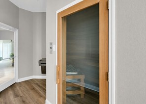 Ein Korridor mit einer Holztür, die zu einem Raum mit einer Glastür, einer Bank und einem kleinen Tisch mit einem Heizgerät führt.