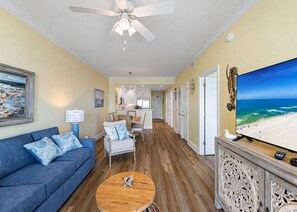 Ein Wohnzimmer mit blauem Sofa, einem Holztisch, einem Deckenventilator, einem Fernseher, der eine Strandansicht zeigt, und einer Küche im Hintergrund.