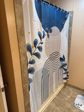 A bathroom with a shower curtain featuring a leaf and wave pattern, a tiled floor, and a showerhead.