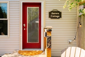 Una puerta roja con un panel de vidrio, un poste de madera con una placa que dice 'Aurora Cottage' y una valla de madera con un accesorio de luz.