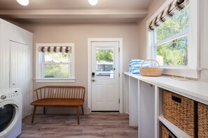 A laundry room with a washing machine, a bench, a door, and a window with a view of trees.
