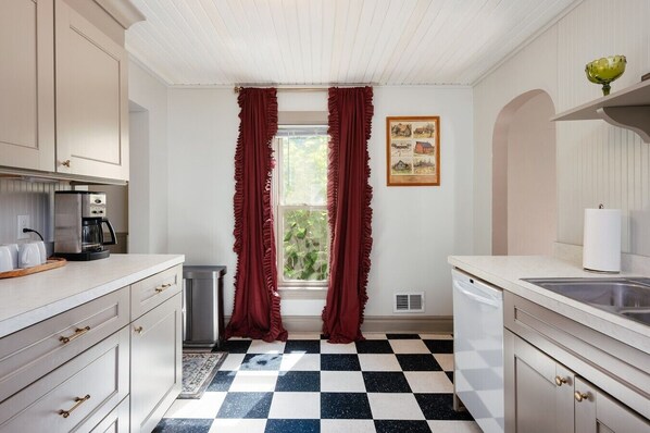 Una cocina con piso a cuadros, una cafetera, un lavabo y un ventanal con cortinas.