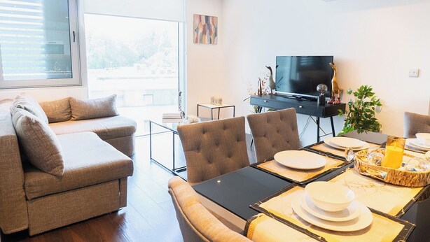 Ein modernes Wohnzimmer mit einer Couch, einem Esstisch, Stühlen, einem Flachbildfernseher und einem großen Fenster.