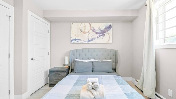 A bedroom with a bed, a nightstand, a lamp, a painting, and a window with curtains. - Elegant Living in a 1-Bedroom Suite (Edmonton)