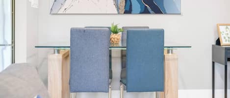 Un comedor moderno con una mesa de vidrio y sillas de metal, un elemento decorativo en la pared y una pequeña planta sobre la mesa.
